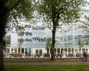 Glazen gevel van het AKN-gebouw in Hilversum met grote zwarte tekst en bomen op de voorgrond.