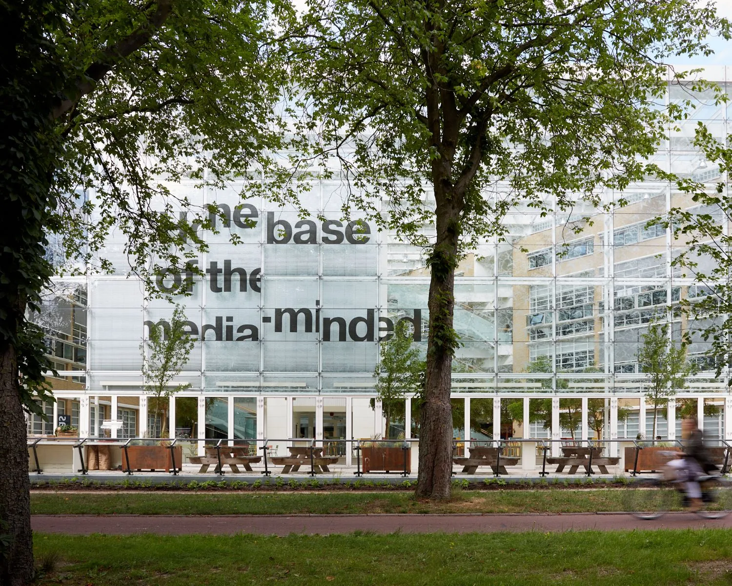Glazen gevel van het AKN-gebouw in Hilversum met grote zwarte tekst en bomen op de voorgrond.
