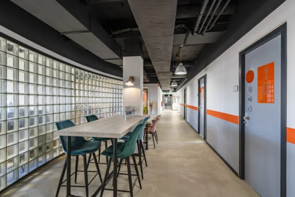 Moderne algemene ruimte in Aalsmeer met glazen bouwstenen, hoge tafels en feloranje accenten langs de deuren en muren.