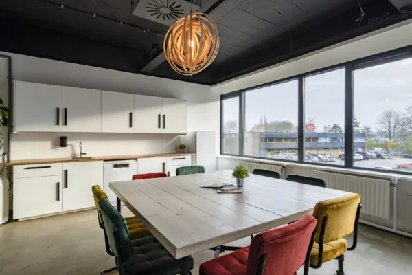 Moderne vergaderruimte in de Aalsmeer-faciliteit met kleurrijke stoelen, een houten tafel, keuken en groot raam met uitzicht op de parkeerplaats.