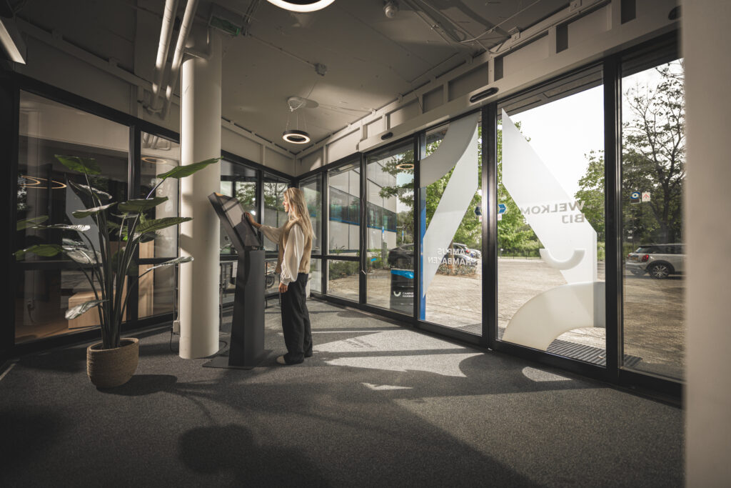 Vrouw meldt zich aan bij een digitale aanmeldzuil in de entree van een modern kantoorpand.