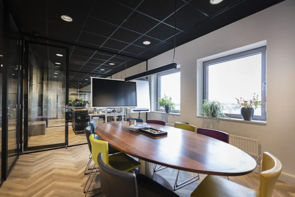 Vergaderruimte in een academie met ovale houten tafel, kleurrijke stoelen, groot scherm en ramen met planten.