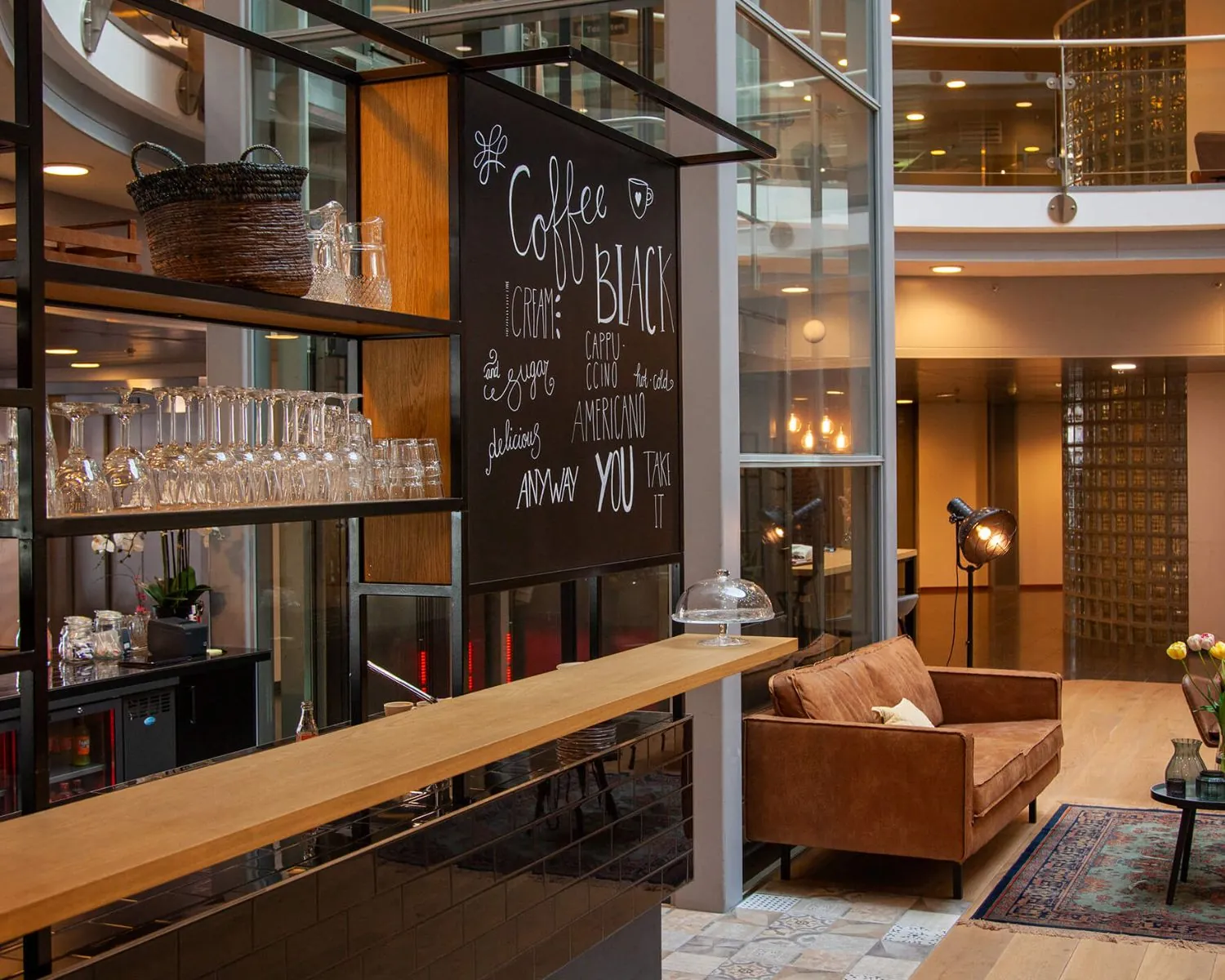 Modern ingerichte koffiehoek met een bruine bank, glazen rek en handgeschreven menubord in een gebouw in Almere.