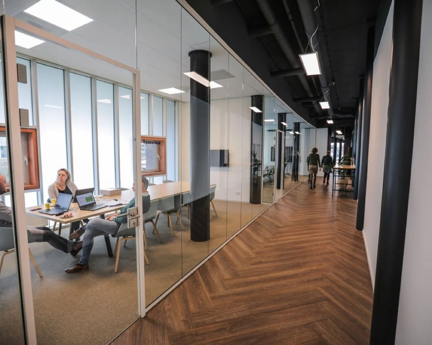 Mensen zitten te vergaderen in een glazen vergaderruimte langs een moderne kantoorhal in Amsterdam-Amersfoort.