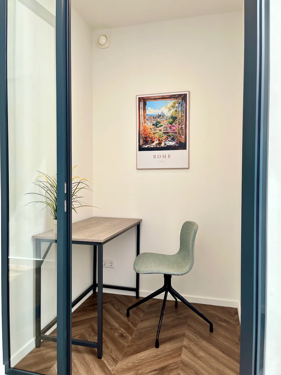 Kleine belruimte met een bureau, stoel, plant en een poster van Rome aan de muur.