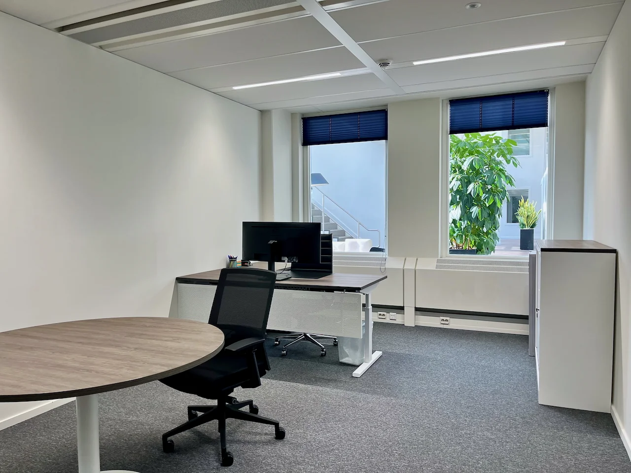 Moderne en lichte kantoorruimte met bureau, vergadertafel en uitzicht op een binnentuin met planten.
