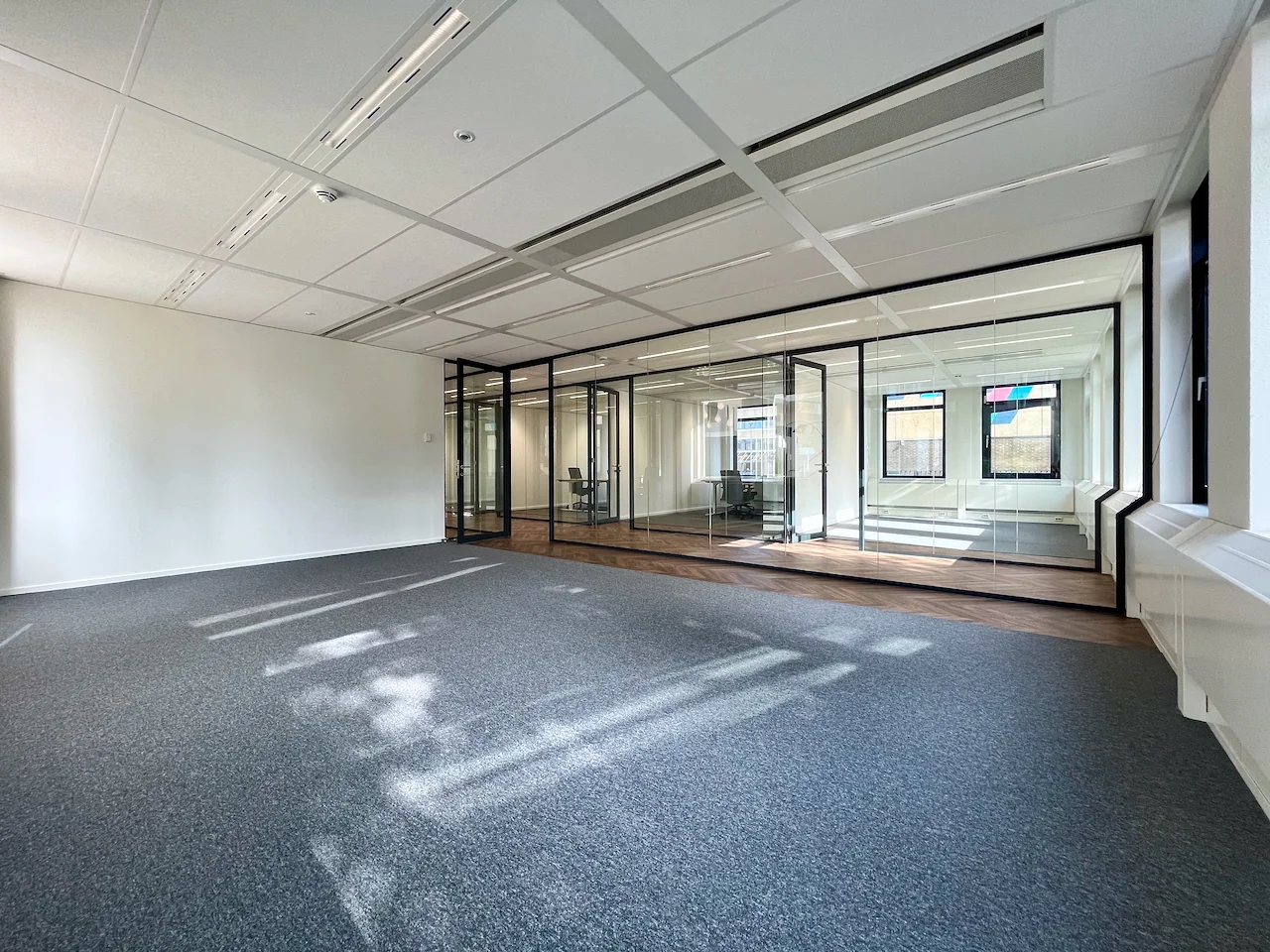 Lichte, lege kantoorruimte met glazen wanden en zicht op een ingerichte vergaderruimte.