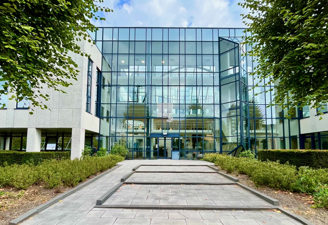 Moderne glazen voorgevel van Gebouw 0.03 met ingang en trappenpartij omringd door groen.