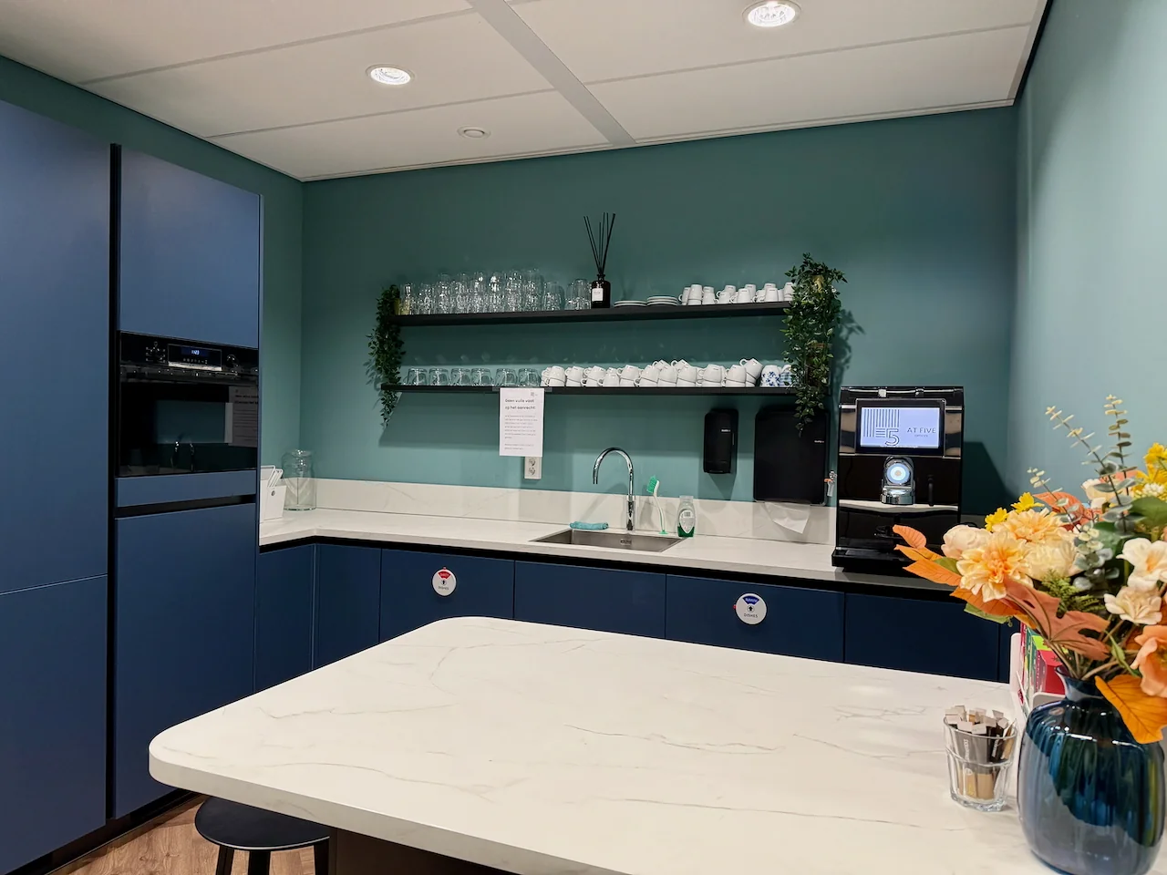 Moderne pantry met blauw keukendesign, koffieautomaat, open planken met servies en een vaas met bloemen op het aanrecht.