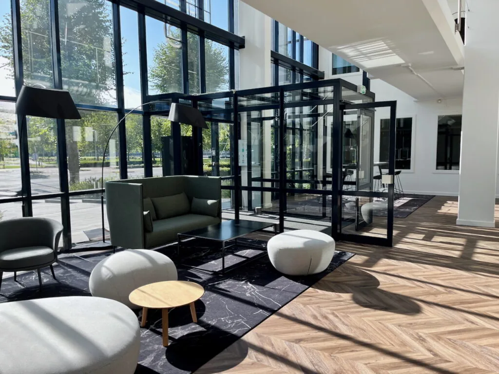 Moderne loungehoek genaamd Lounge 0.04 met designmeubels, veel daglicht en glazen wanden in een stijlvolle kantoorruimte.