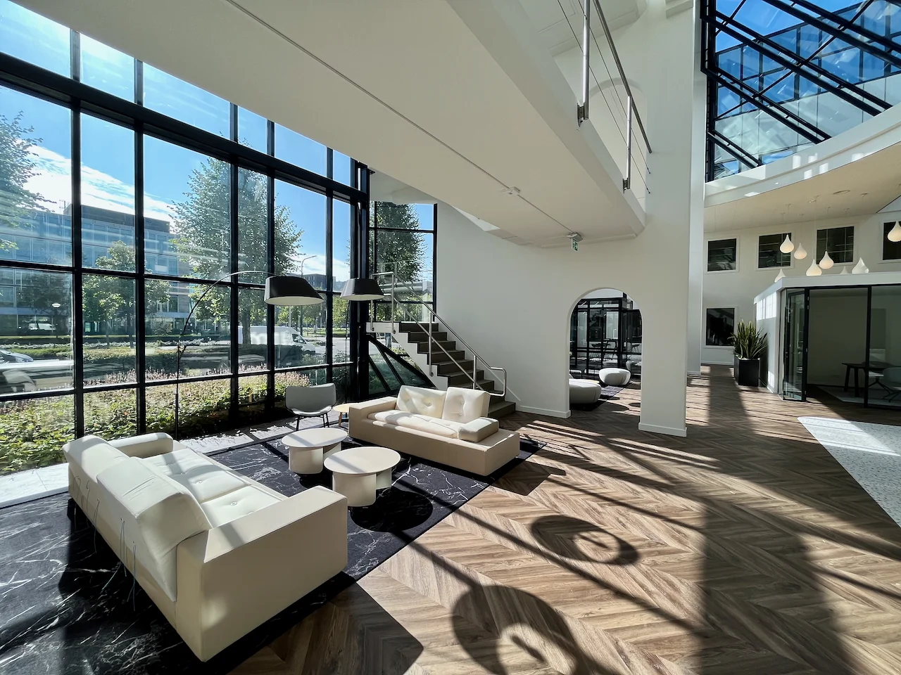 Lichte en moderne lounge 0.05 met witte banken, grote ramen en houten vloer in een open kantoorruimte.