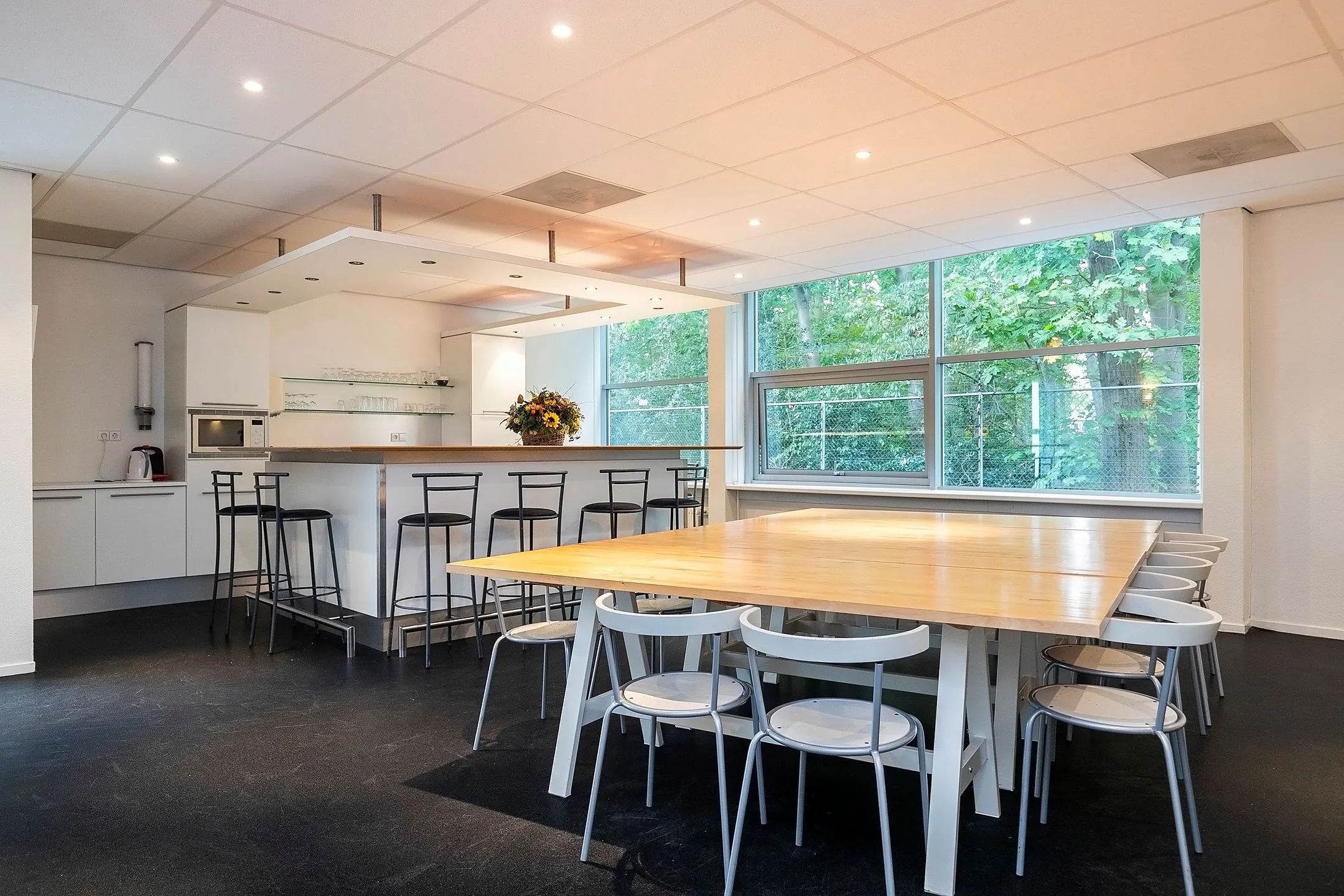 Moderne gemeenschappelijke ruimte aan de Banningstraat 3 met een open keuken, bar en grote eettafel bij een raam met uitzicht op groen.