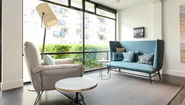 Moderne wachtruimte met grote ramen, een grijze fauteuil, een blauwe bank en een vloerkleed aan de Bezuidenhoutseweg 161.