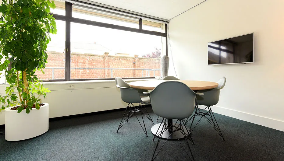 Vergaderruimte met ronde tafel, vijf stoelen, een flatscreen aan de muur en een grote plant bij het raam aan de Bezuidenhoutseweg 161.