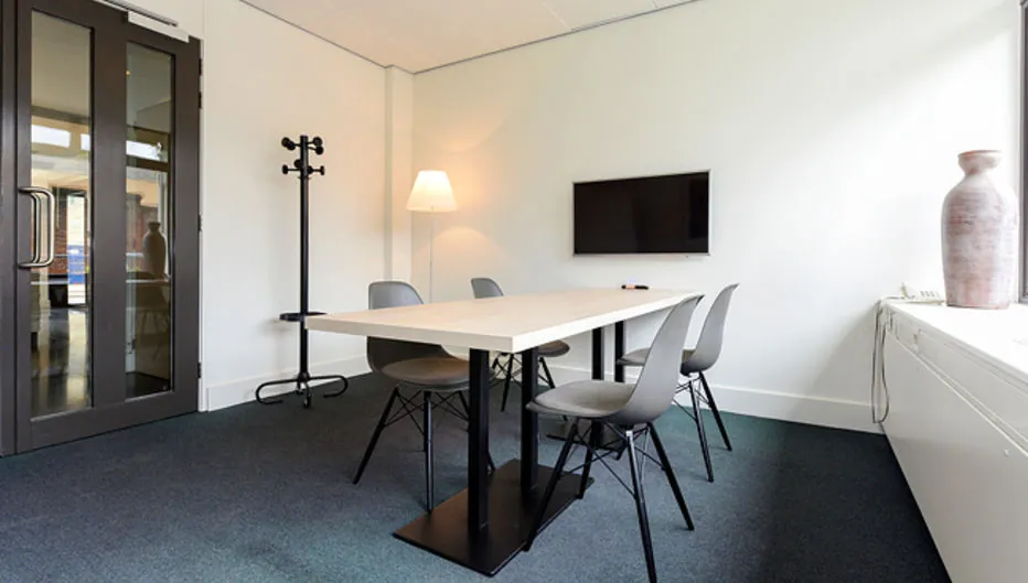 Vergaderruimte aan de Bezuidenhoutseweg 161 met een tafel, zes stoelen, een flatscreen, kapstok en staande lamp.