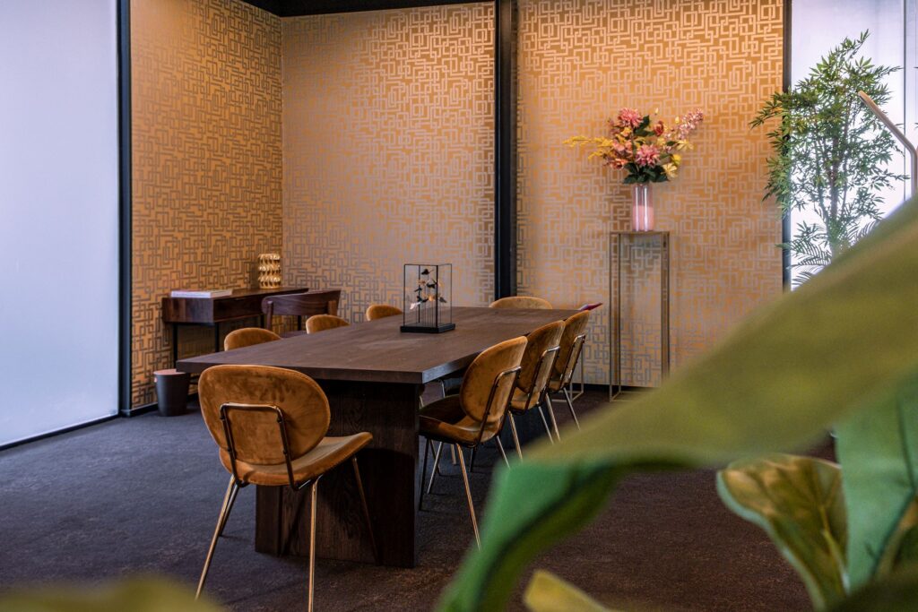 Vergaderruimte aan de Ceintuurbaan 28 met houten tafel, fluwelen stoelen en decoratieve elementen zoals bloemen en planten.