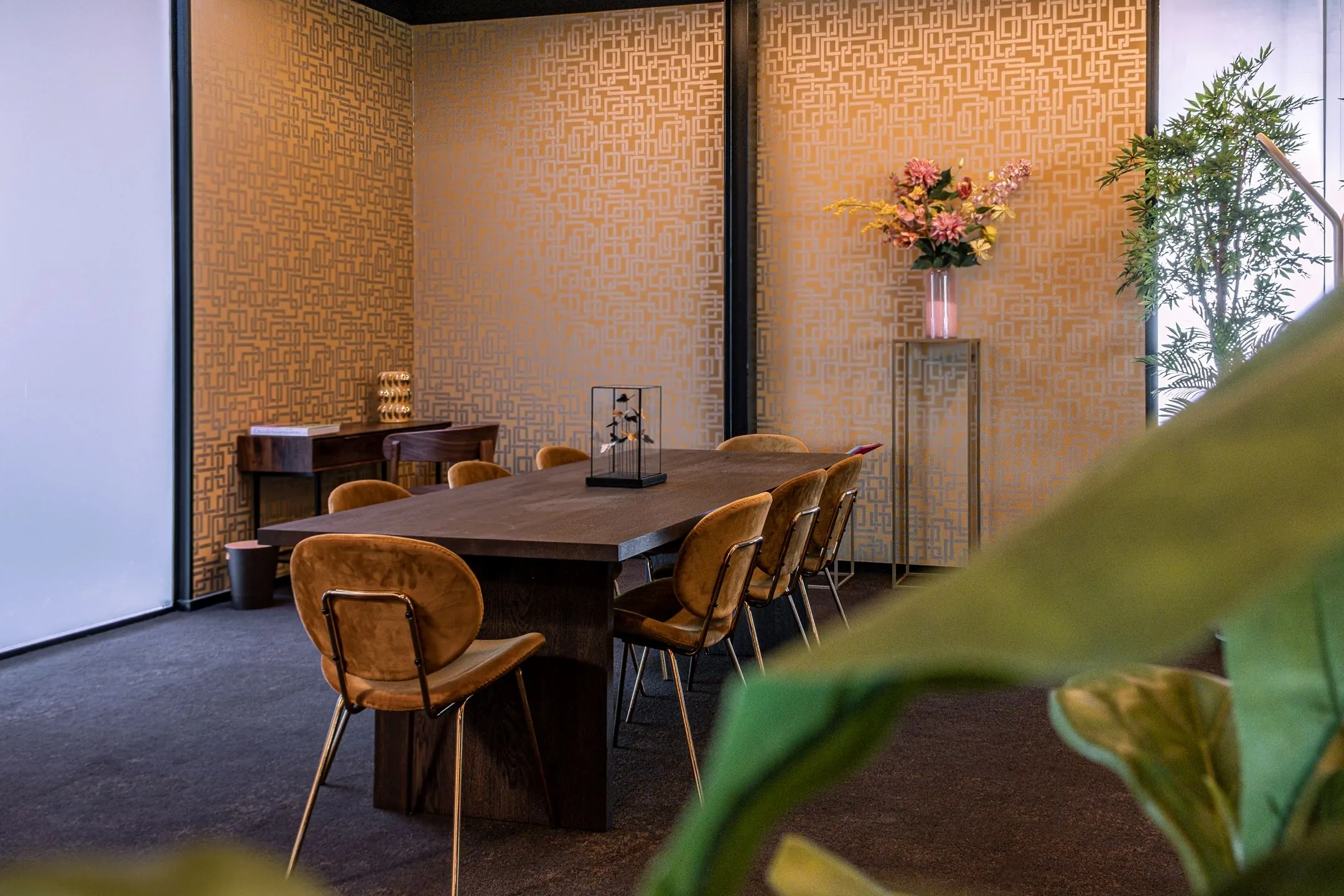Vergaderruimte aan de Ceintuurbaan 28 met houten tafel, fluwelen stoelen en decoratieve elementen zoals bloemen en planten.
