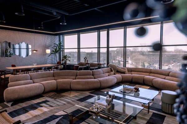 Modern ingerichte lounge op Ceintuurbaan 28 met grote ronde bank, glazen salontafels en uitzicht op stad via hoge ramen.