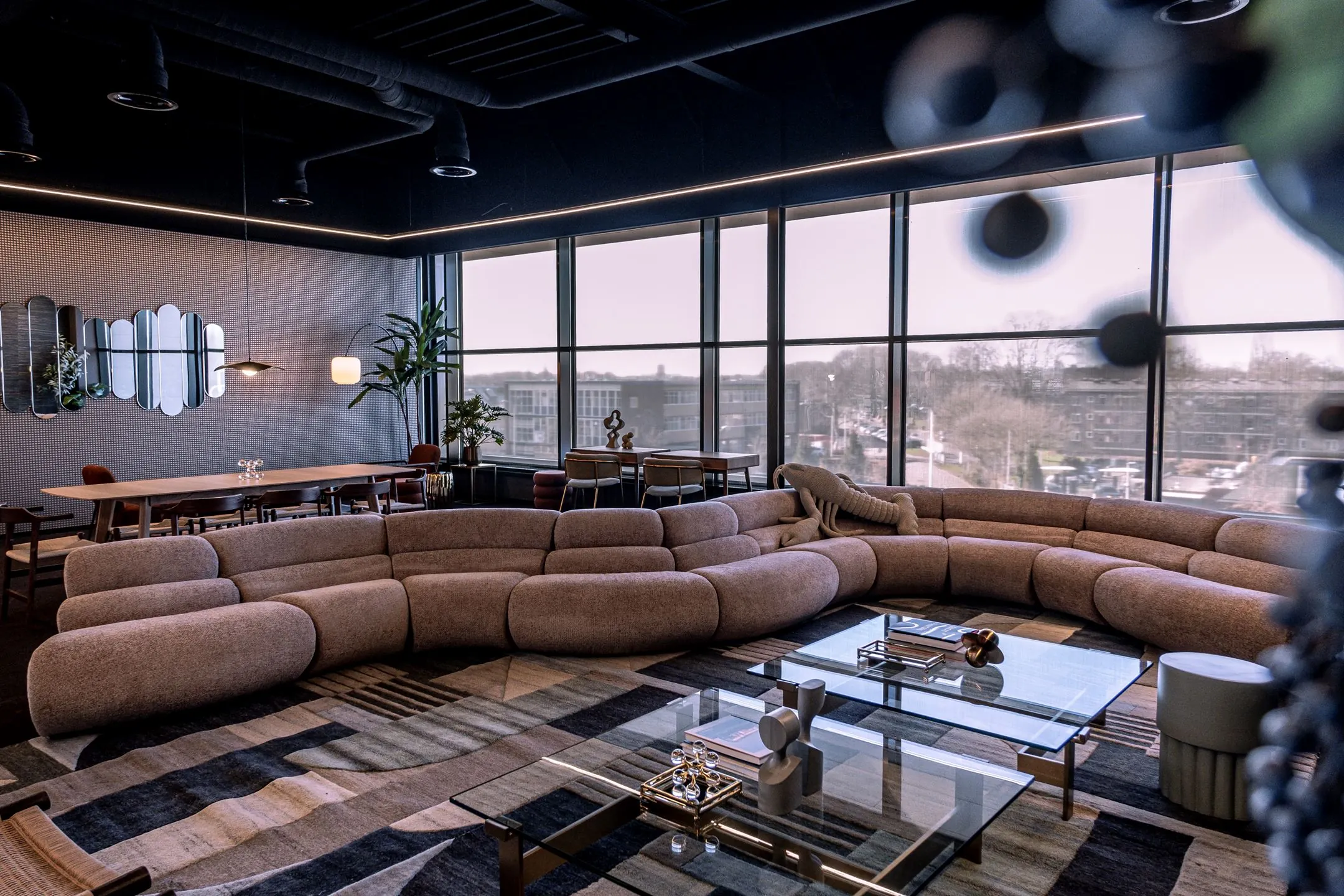 Modern ingerichte lounge op Ceintuurbaan 28 met grote ronde bank, glazen salontafels en uitzicht op stad via hoge ramen.
