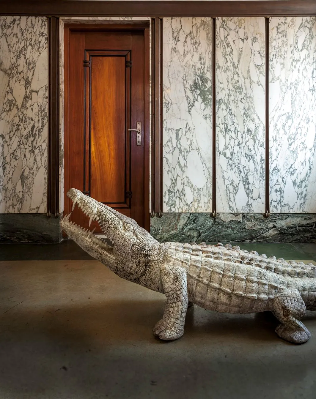 Beeld van een marmeren hal met houten deur en een decoratief krokodilbeeld op de vloer.