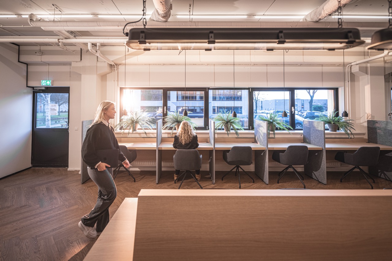 Moderne co-working space in de Delta bibliotheek met werkplekken langs het raam en twee personen aan het werk.