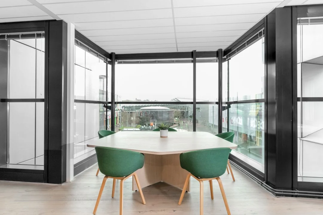 Vergaderruimte met een houten tafel, groene stoelen en uitzicht op de Coolsingel in Rotterdam door grote glazen ramen.