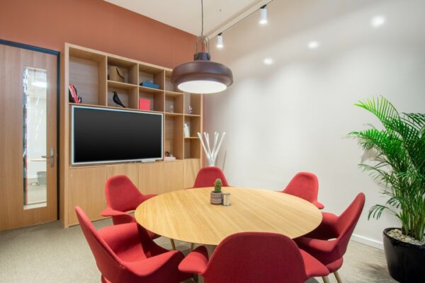 Vergaderruimte met ronde houten tafel, rode stoelen, wandkast met tv en decoratie, en een grote plant in de hoek.