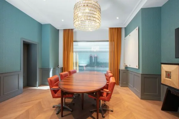 Vergaderruimte in Coolsingel 65 met ovale houten tafel, oranje stoelen, grote kroonluchter en hoge ramen met okergele gordijnen.
