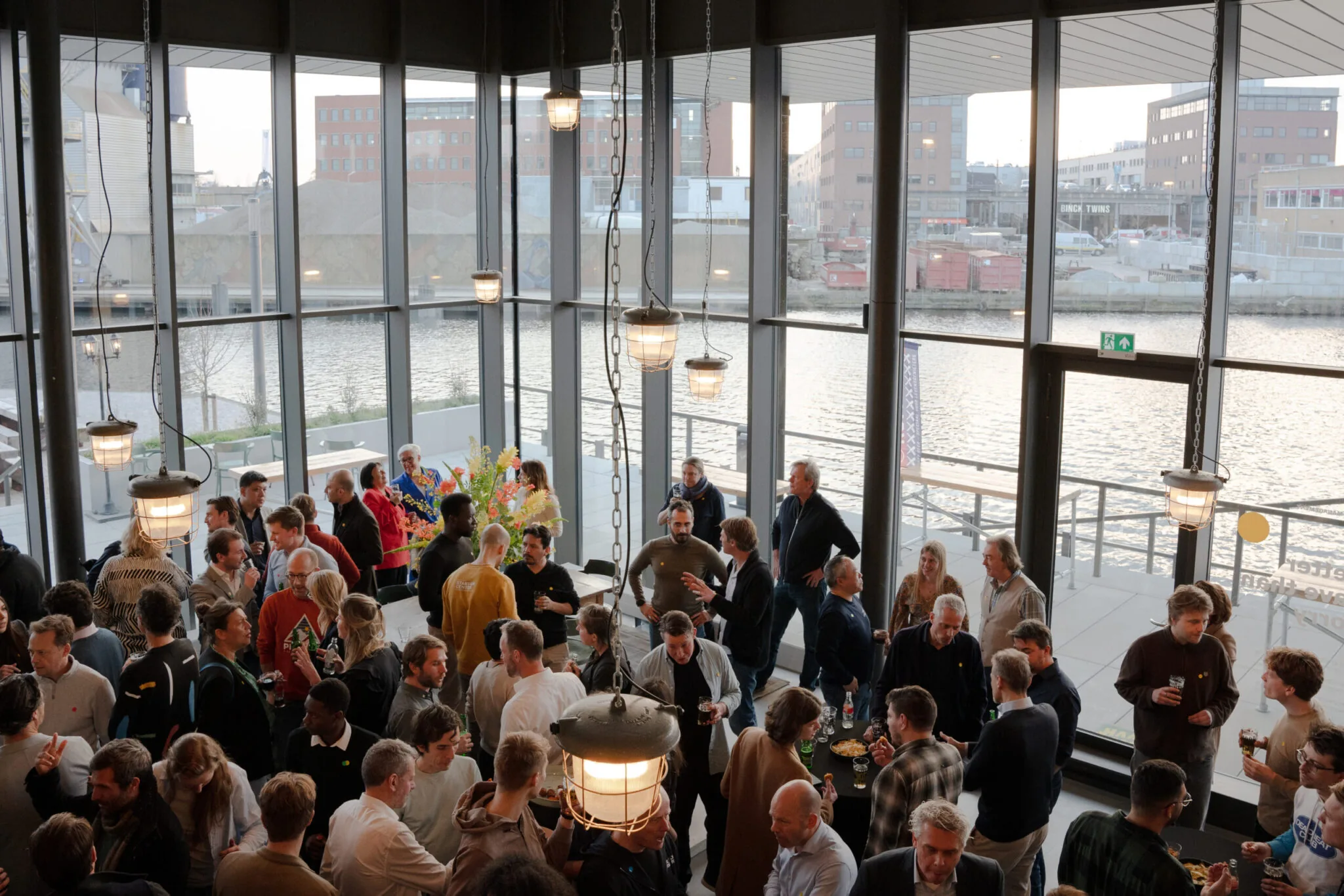 Grote groep mensen socialiseert tijdens een evenement in een moderne ruimte met uitzicht op het water.
