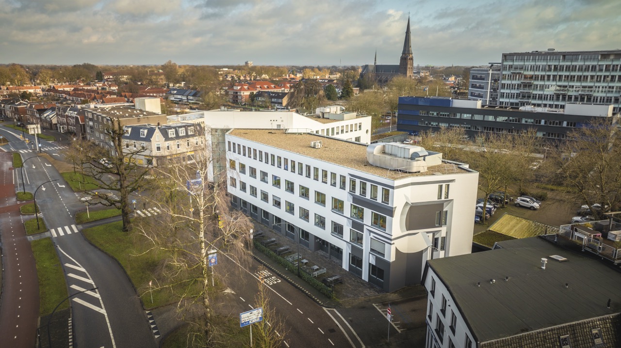 Luchtfoto van een modern wit kantoorgebouw aan een kruispunt in een stedelijke omgeving met kerktoren op de achtergrond.