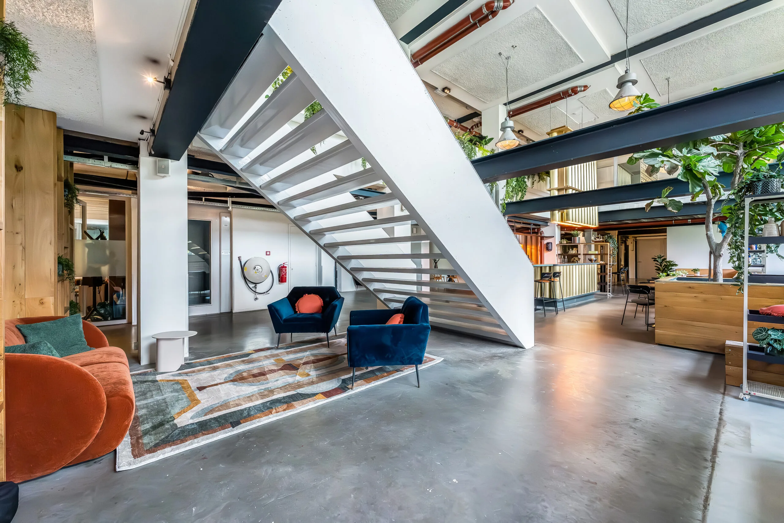 Moderne open kantoorruimte met industriële elementen, kleurrijke zithoek en centrale witte trap.