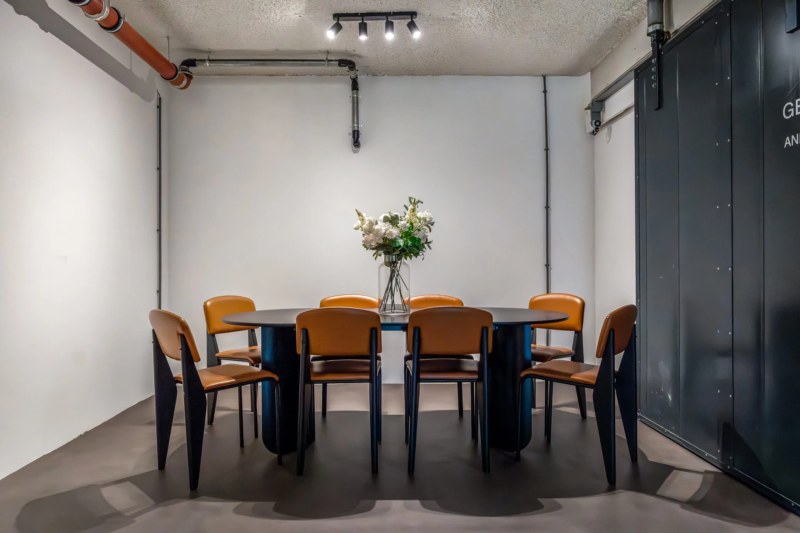 Vergaderruimte met een ovaal zwarte tafel, omringd door zes bruine stoelen en een vaas met bloemen in het midden.