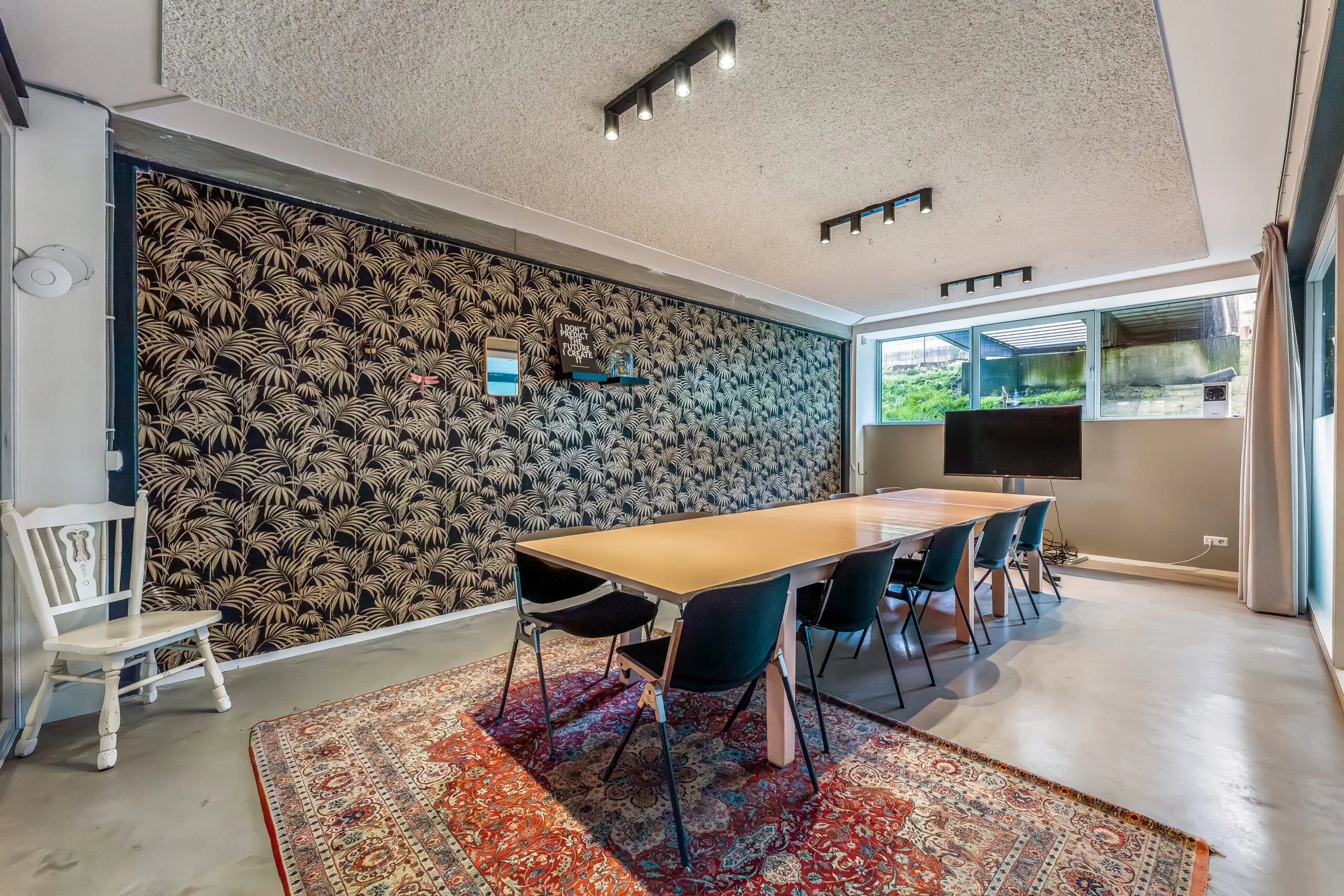 Moderne vergaderruimte met een lange tafel, zwarte stoelen, een sierkleed en een accentmuur met palmenpatroon.