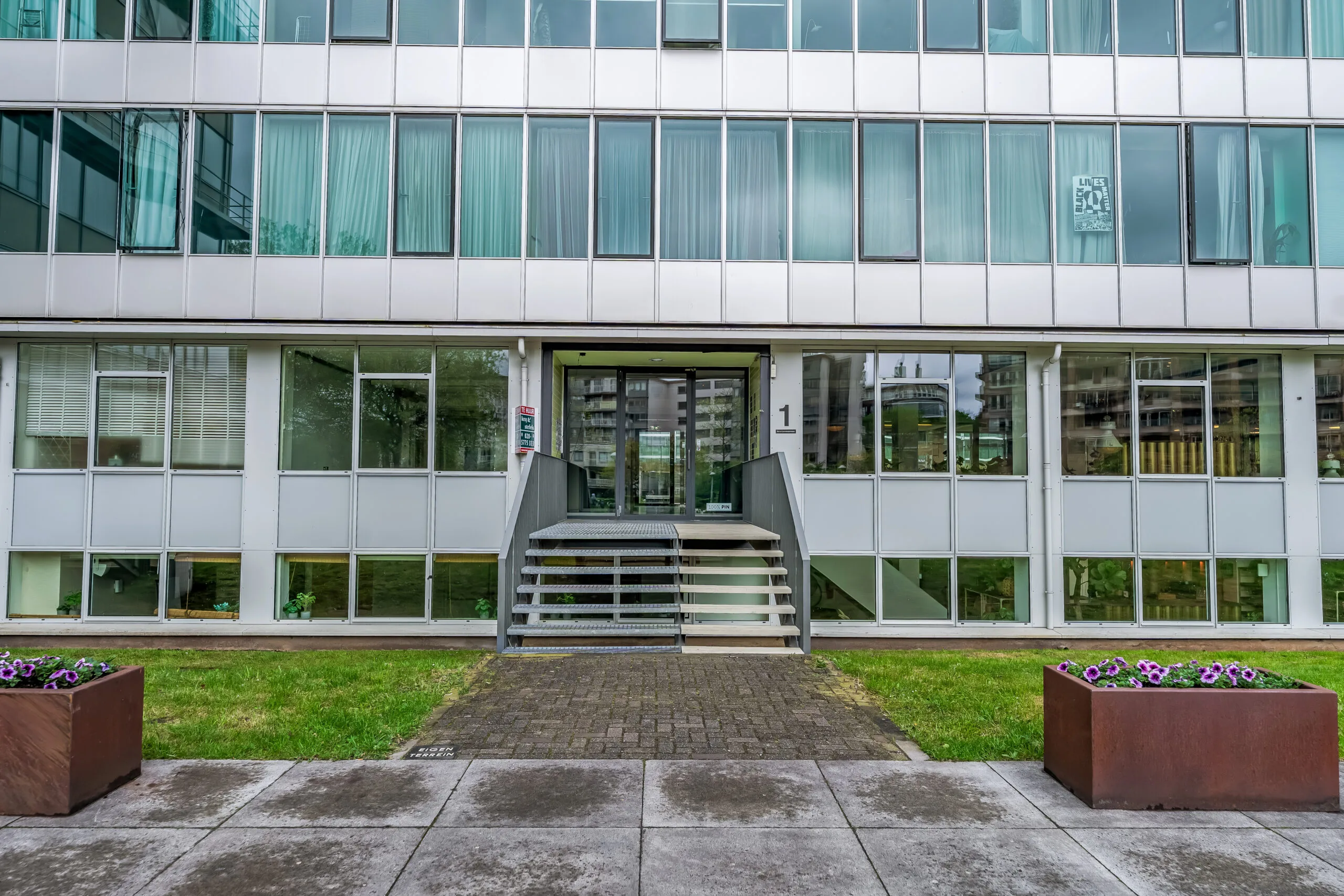 Vooraanzicht van een modern kantoorgebouw met glazen gevel en een centrale entree met trap en hellingbaan.