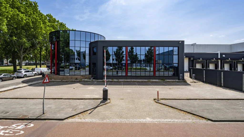 Modern kantoorpand met spiegelende gevel en laadperrons, gezien vanaf een parkeerterrein.
