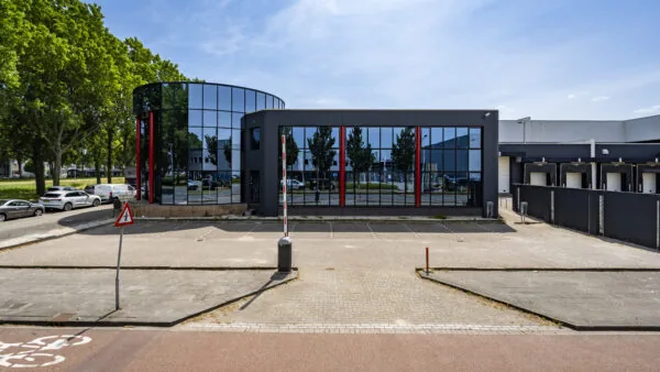 Modern kantoorpand met spiegelende gevel en laadperrons, gezien vanaf een parkeerterrein.