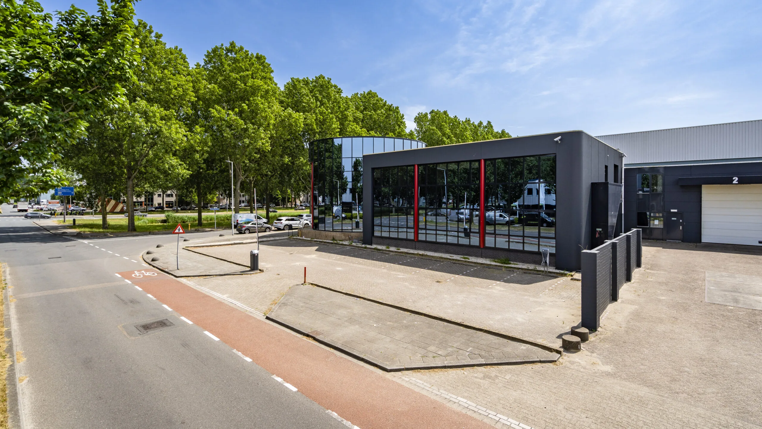 Modern bedrijfsgebouw met grote spiegelramen aan een rustige straat met een fietspad en veel groen.