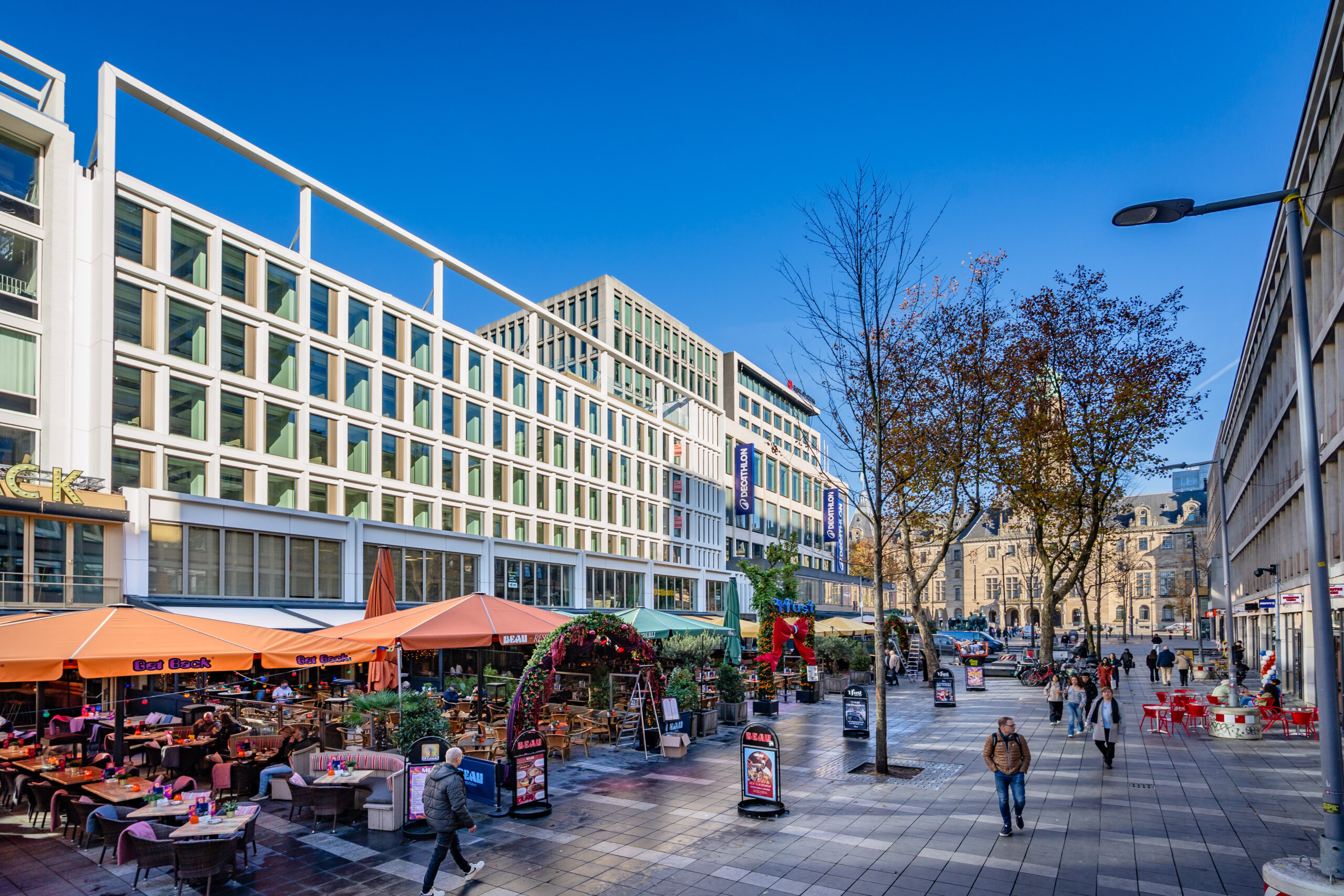 Stadsplein met moderne gebouwen, terrassen en voorbijgangers onder een heldere blauwe lucht.