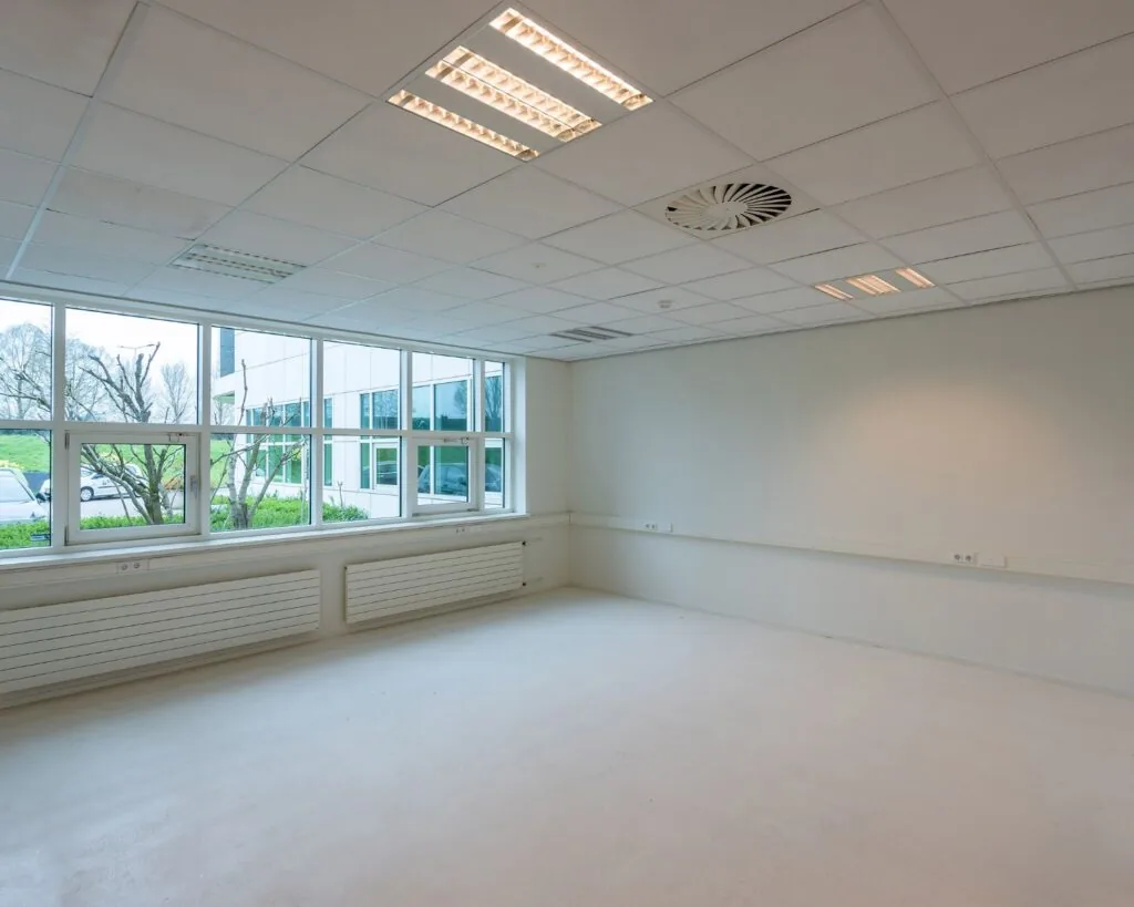 Lege, lichte kantoorruimte met systeemplafond, grote ramen en witte muren.