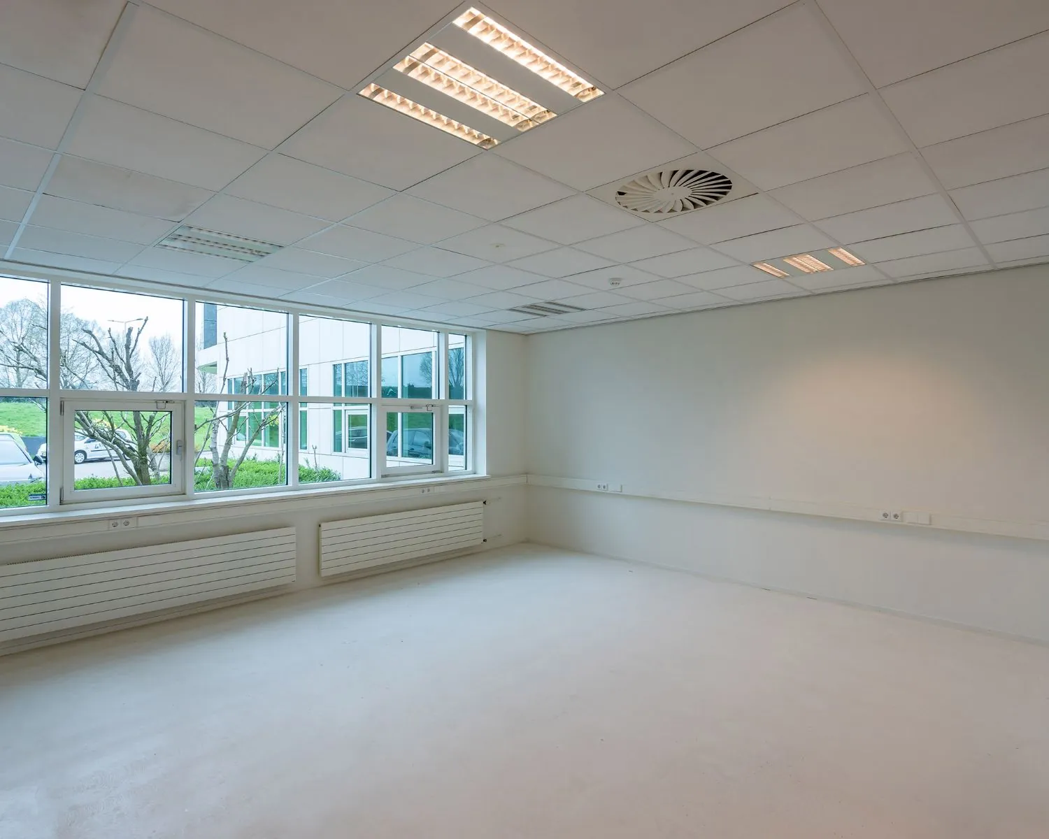 Lege, lichte kantoorruimte met systeemplafond, grote ramen en witte muren.
