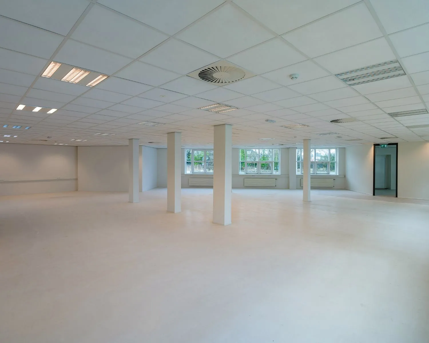 Lege, ruime en lichte kantoorruimte met witte muren, plafondplaten en grote ramen.