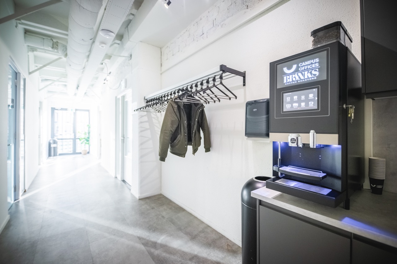 Lichte kantoorhal met een koffieautomaat van Campus Offices BRNKS, een kapstok met jassen en hangers.