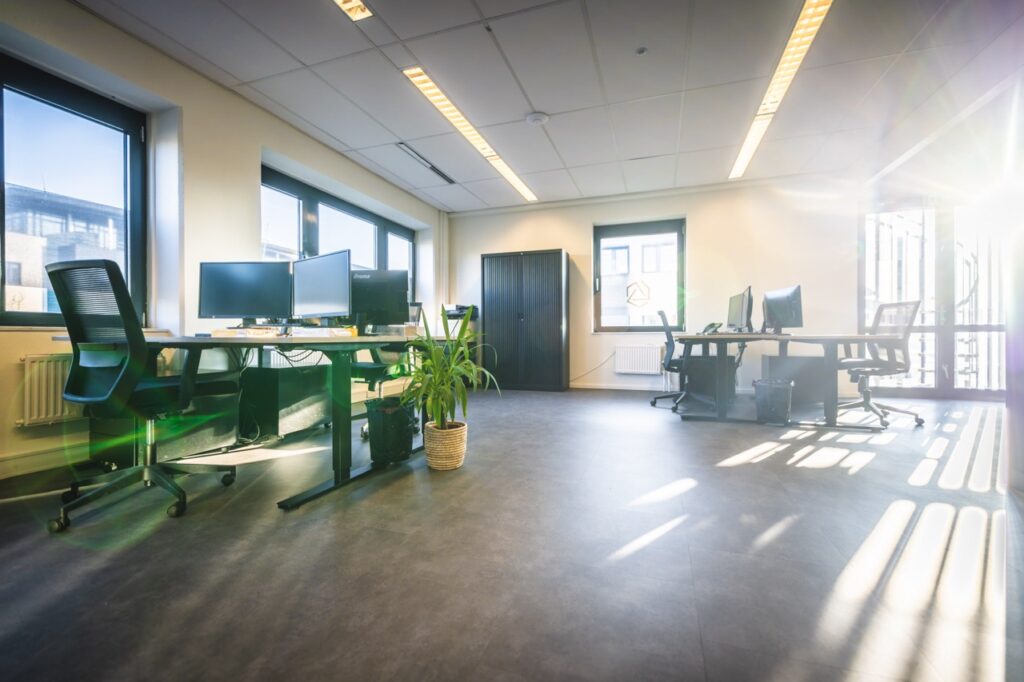 Lichte en moderne kantoorruimte met bureaus, bureaustoelen, computerschermen en grote ramen waar zonlicht naar binnen schijnt.