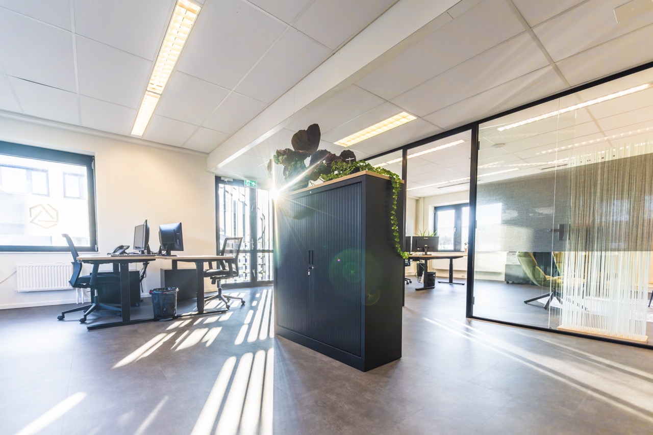 Modern en licht kantoorinterieur met bureaus, glazen wanden en een zwarte kast met planten.