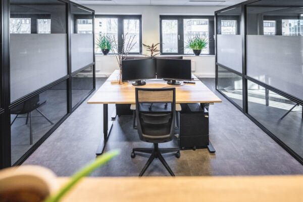 Modern kantoor met een houten bureau, twee computerschermen, een bureaustoel en planten bij het raam tussen glazen scheidingswanden.