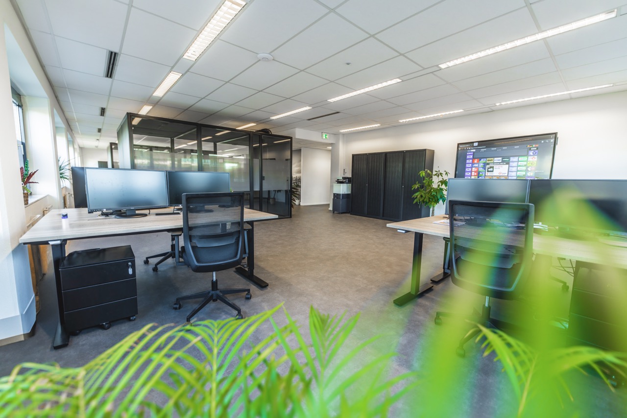 Moderne open kantoorruimte met bureaus, computerschermen, glazen vergaderruimtes en groene planten.