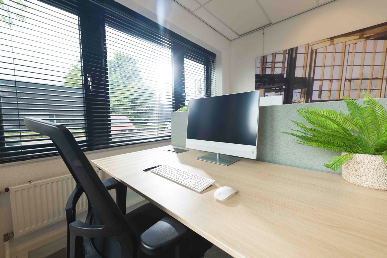 Modern en licht kantoorwerkplek met een computerscherm, toetsenbord, bureaustoel en een plant naast een raam met jaloezieën.