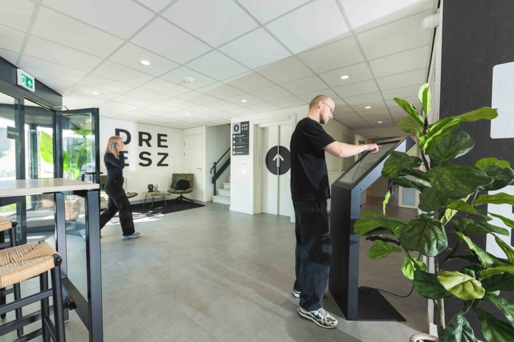 Man gebruikt een touchscreen-zuil in de moderne ontvangstruimte van DREESZ, terwijl een vrouw bellend voorbijloopt.