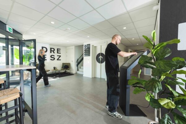 Man gebruikt een touchscreen-zuil in de moderne ontvangstruimte van DREESZ, terwijl een vrouw bellend voorbijloopt.
