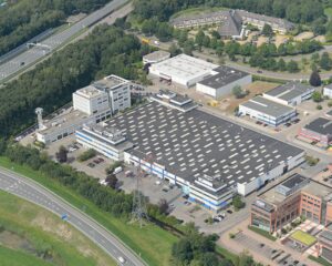 Luchtfoto van een groot industrieel complex met kantoren en parkeerterreinen naast een snelweg in een groene omgeving.